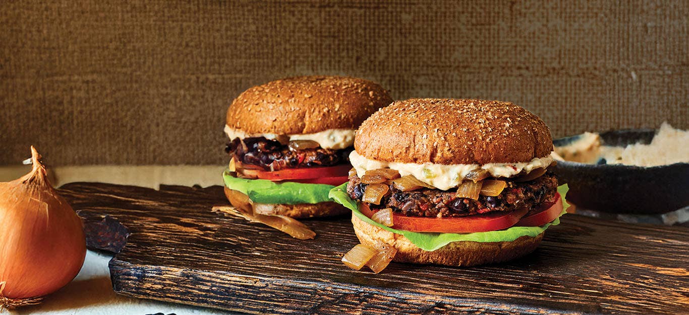 Two Black Bean Smash Burgers with Caramelized Onions on a wooden chopped board with an onion to the left of the board and a small bowl of sauce to the right