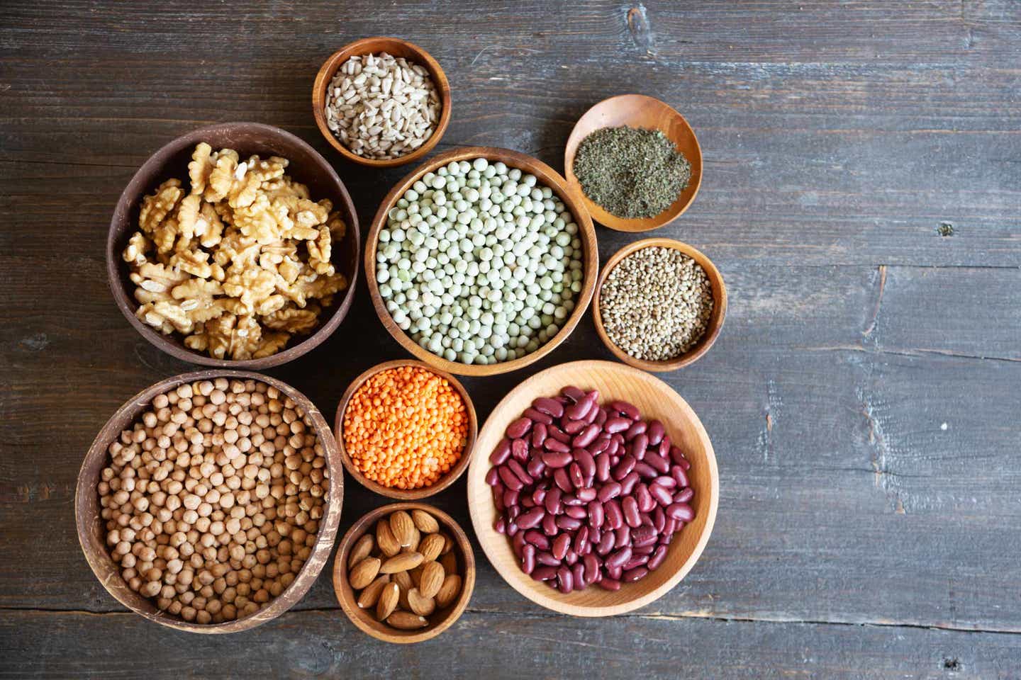 A collection of small and medium size bowls, each filled with legumes or nuts, on a wooden gray background