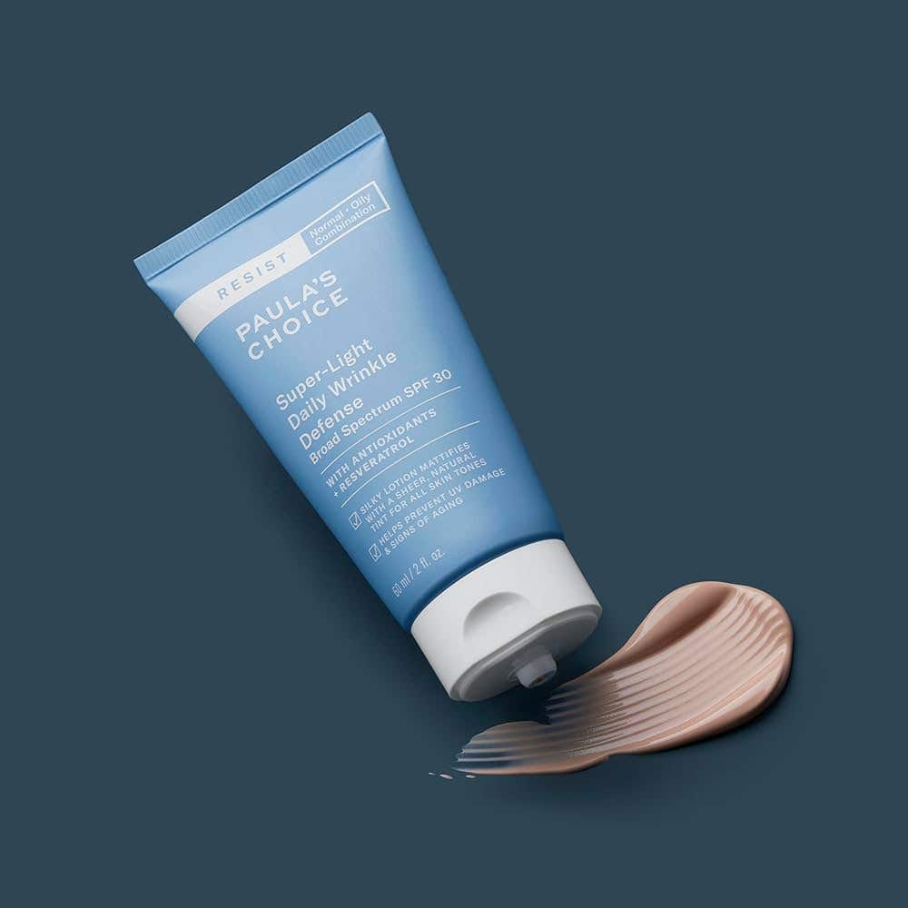 A bottle of Paula's Choice Super Light Daily Wrinkle Defense SPF on a dark blue background, with some of the tinted sunscreen smudged beneath the open cap