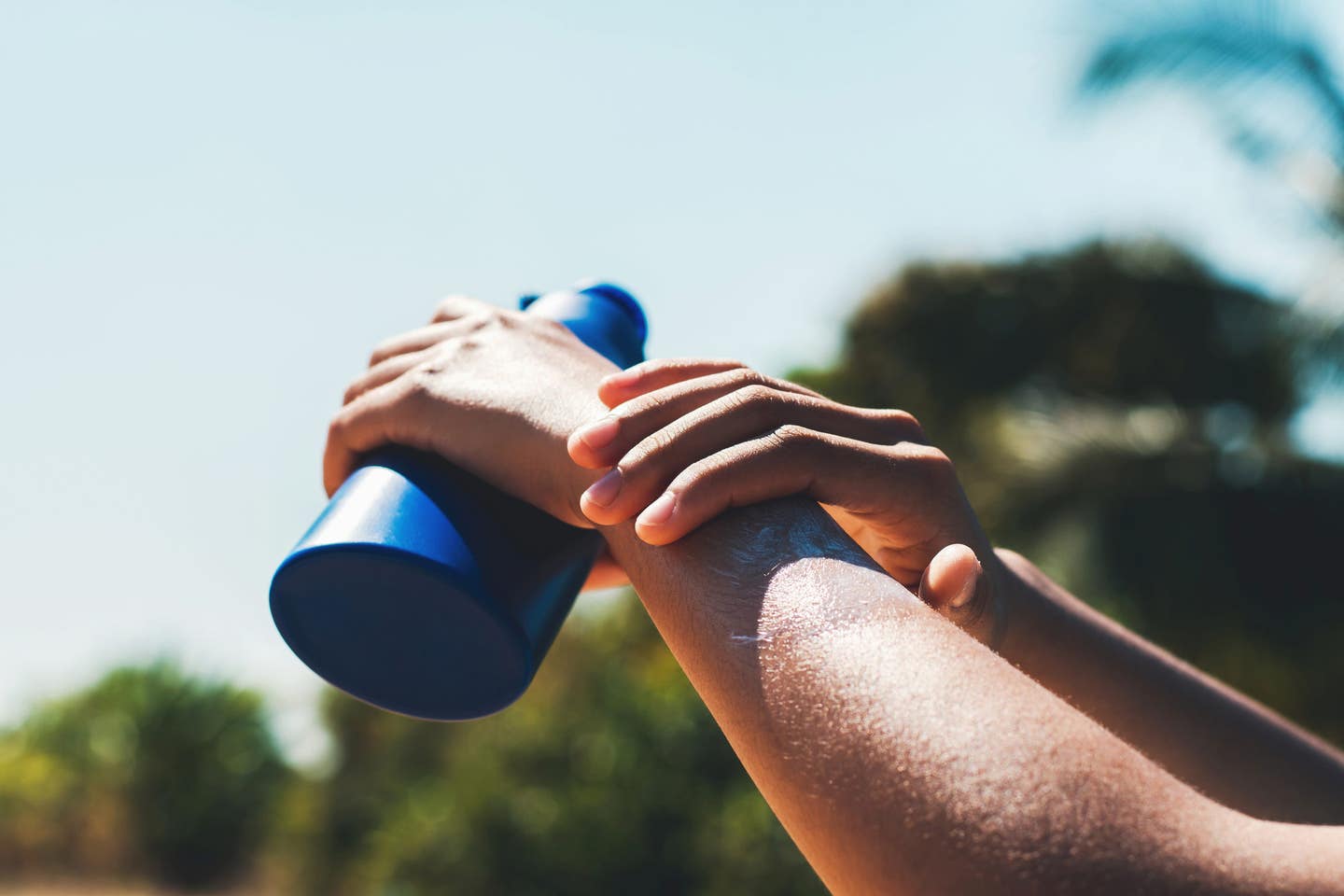 the arms of someone applying sunblock outside, holding the blue bottle of sunblock