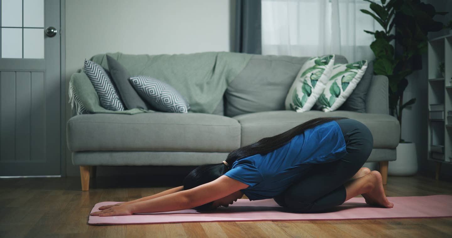 A woman in her dim living room at dusk engaged in child's pose, a restorative yoga pose to help with sleep and relaxation