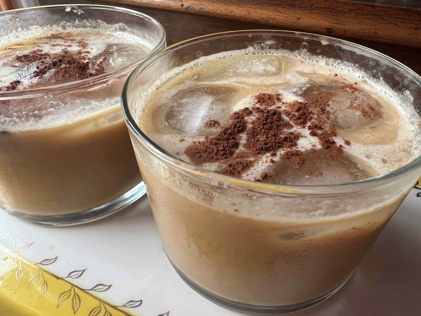 Two glasses of vegan White Russian mocktail lattes sitting on a white and yellow table near a windowsill
