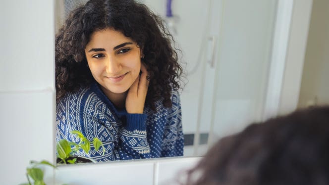 getting-out-of-survival-mode A young woman with curly hair looks into the bathroom mirror and smiles