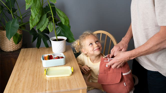 lunchbox a little girl sits at a kitchen table as her did packs her lunchbox full of healthy foods, including strawberries and carrots